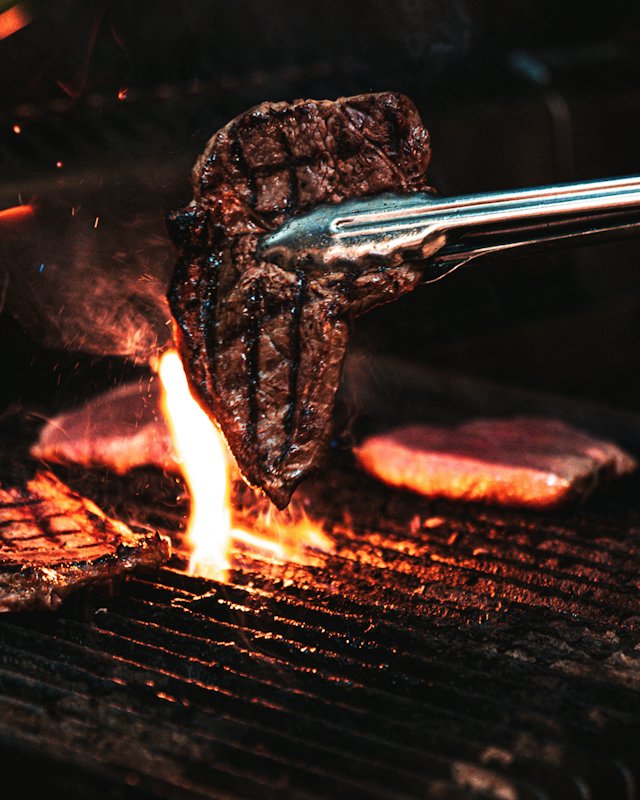 Een tang tilt een gegrild stuk vlees van een vlammende houtskoolgrill